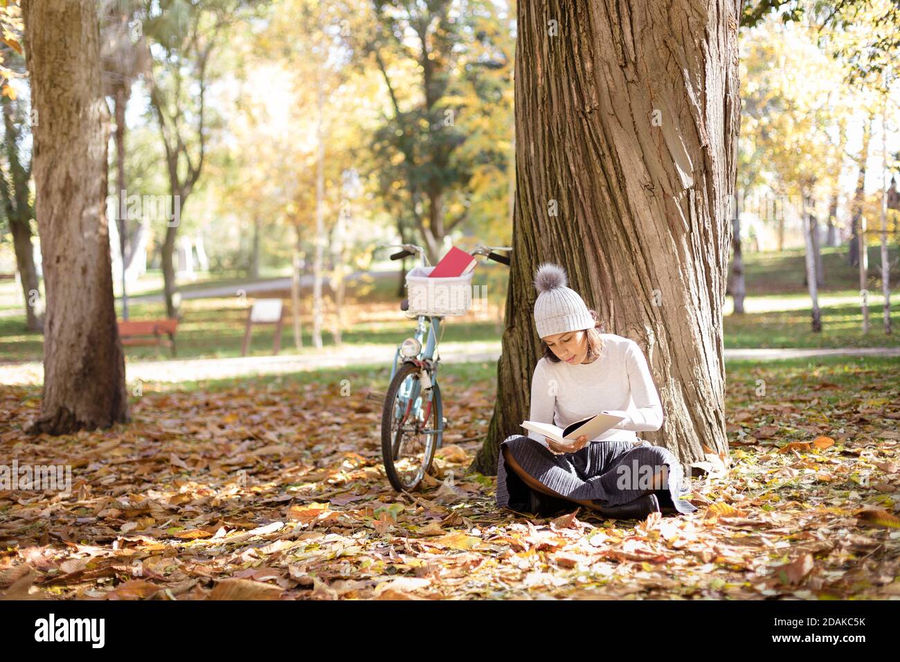 Giovane donna che legge un libro nel parco accanto alla sua bicicletta. È seduta sul terreno circondata da foglie cadute dagli alberi. Spazio per il testo. Foto Stock