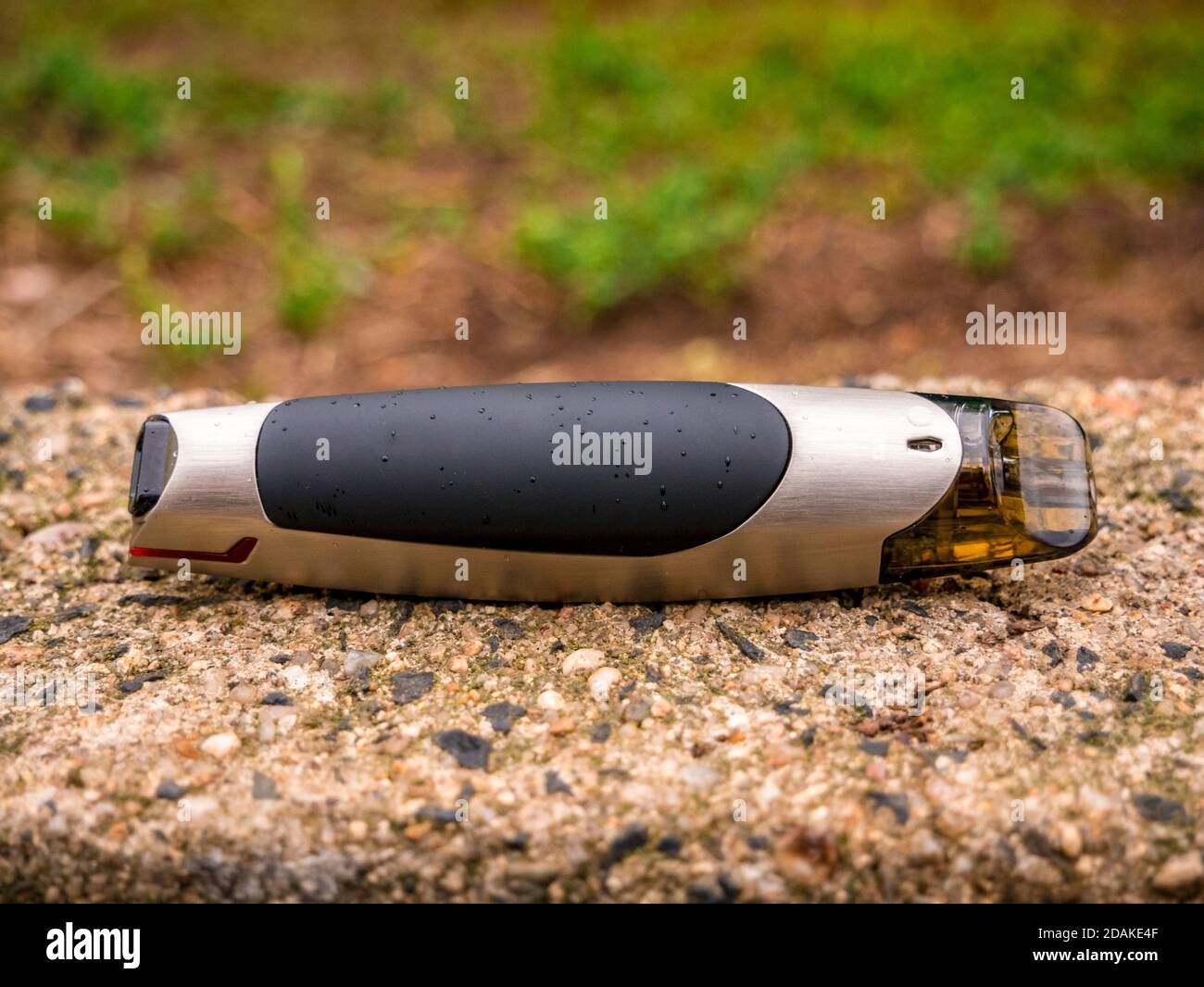 Sigaretta elettronica - il cosiddetto sistema POD per la vaporizzazione,  leggermente macchiato con acqua giacente su un blocco di cemento con verdi  sfocati sullo sfondo Foto stock