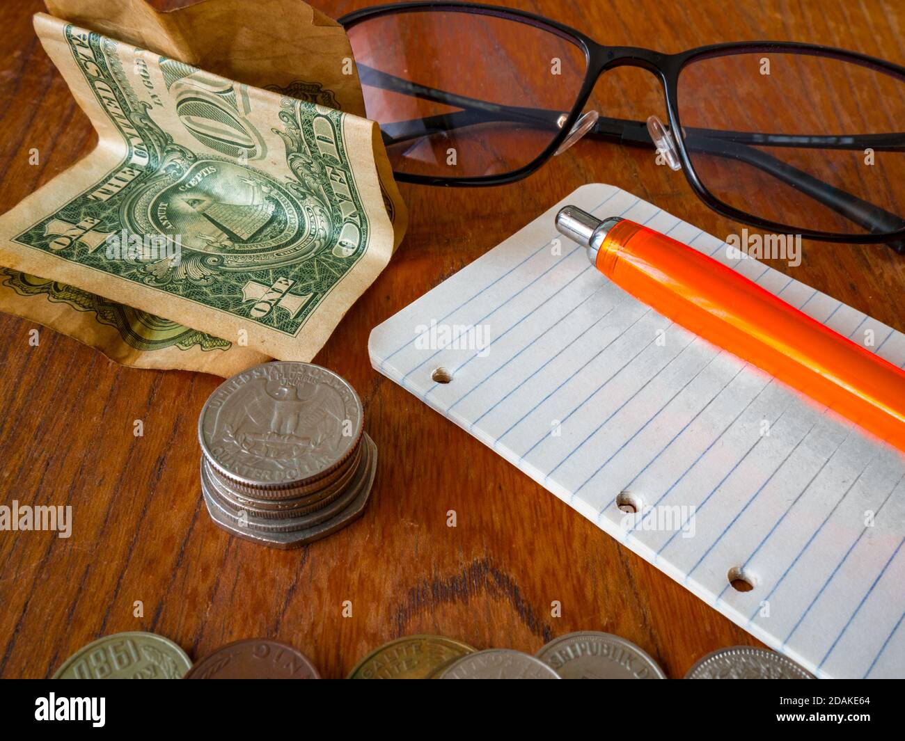 Ha piegato una banconota da dollaro USA, una pila di monete (dollaro del quarto americano sulla parte superiore), un blocco note con una penna, bicchieri e una pila di monete in primo piano. Foto Stock