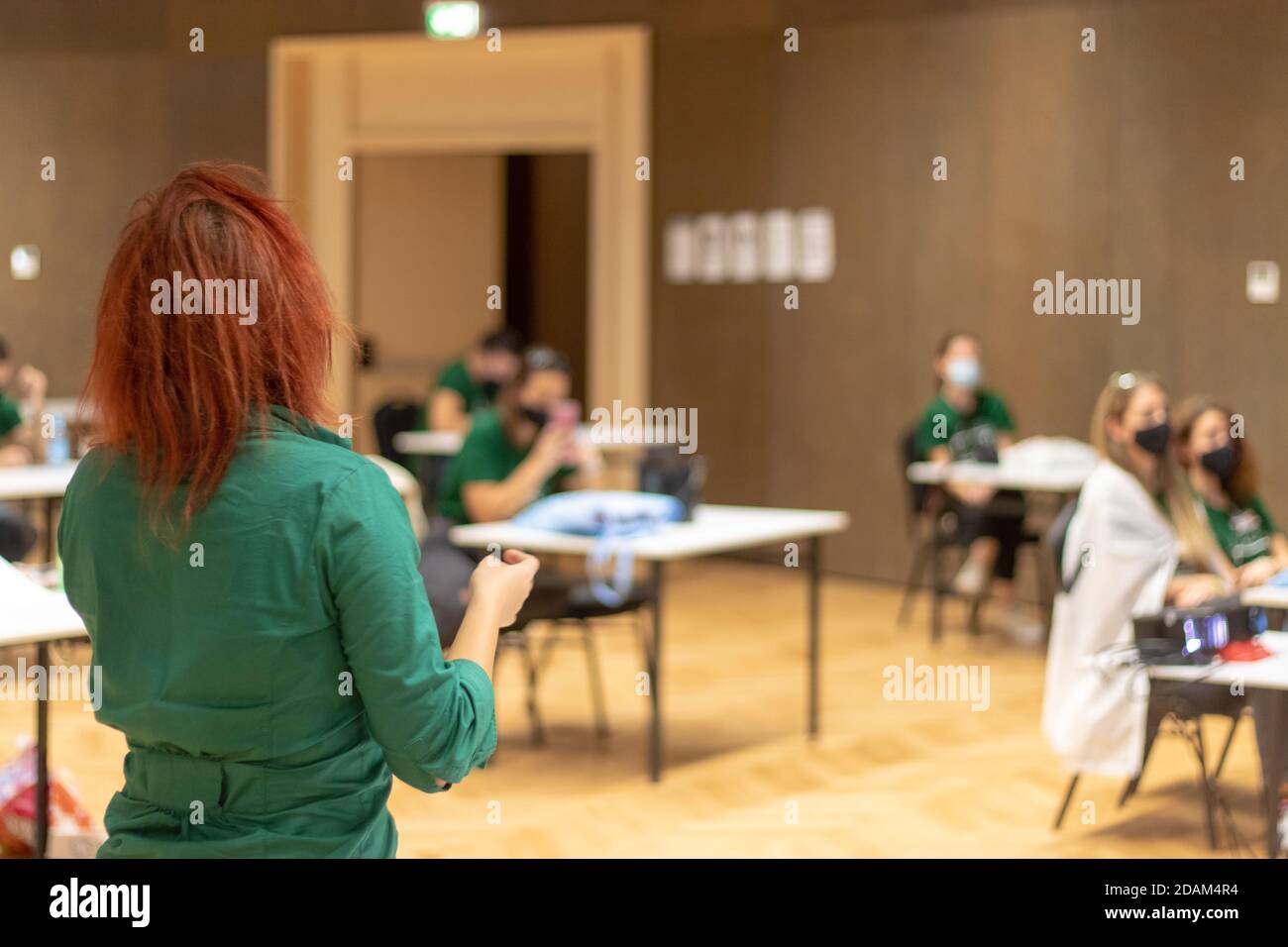 Vista posteriore di una donna irriconoscibile con i capelli rossi che tengono una presentazione all'interno, le persone all'interno seduta con spazio tra di loro, seguendo covid g Foto Stock