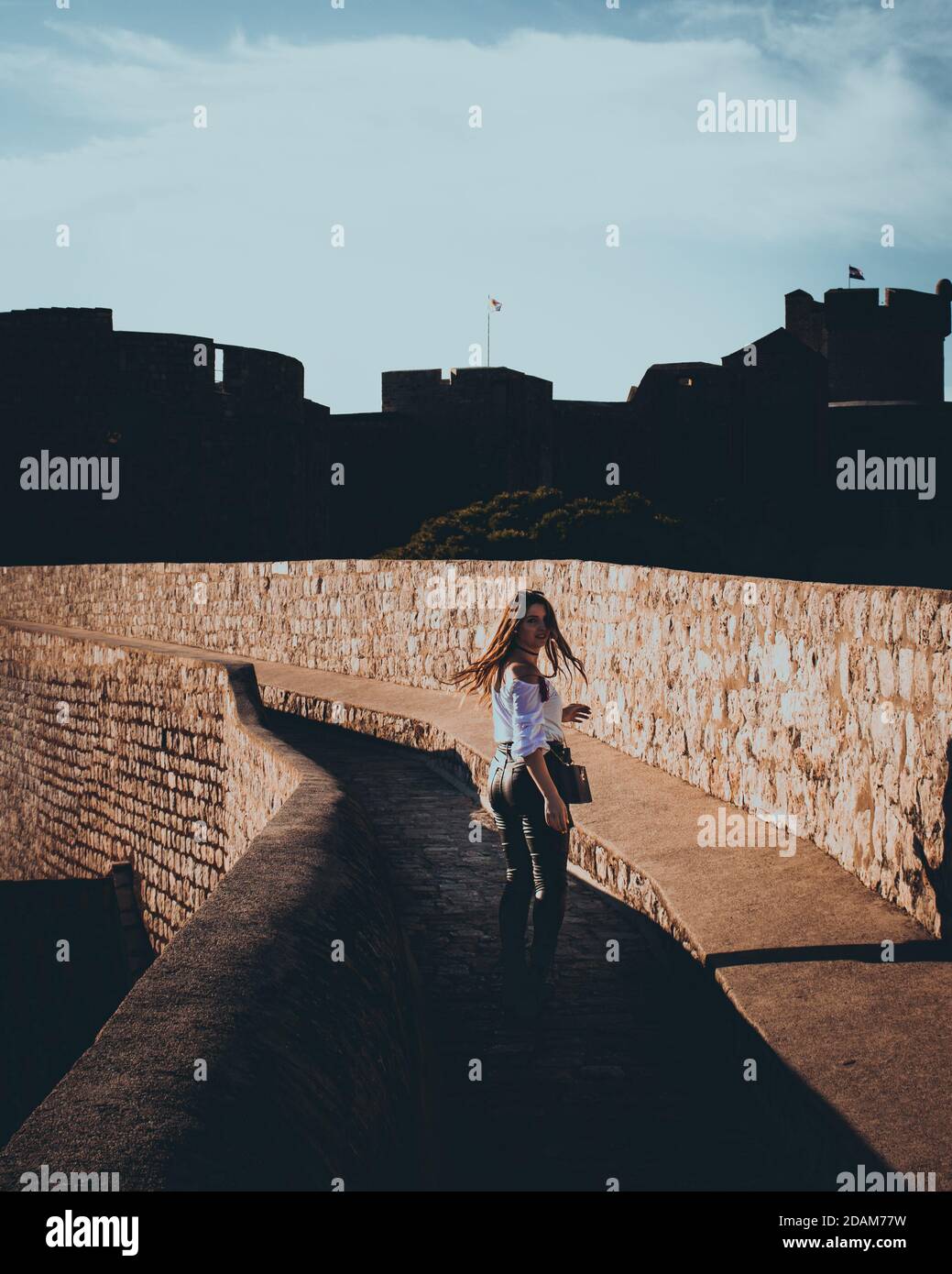 Foto verticale e vista posteriore di una ragazza attraente che cammina lungo le mura della città di Dubrovnik. Modifica della moodia scura con evidenziazioni in bianco e nero Foto Stock