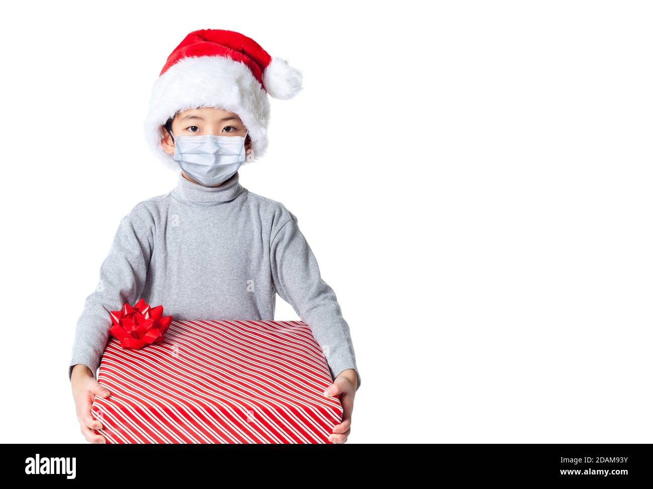 Bambino che riceve regalo di Natale durante il Natale di Covid che indossa il cappello di Santa e la maschera del viso a causa della pandemia, isolato su sfondo bianco con spazio di copia. Foto Stock