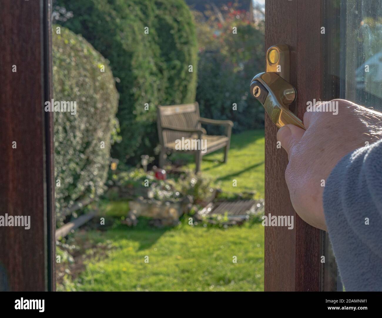 Primo piano POV colpo di una mano di un uomo che tiene una maniglia su una finestra al piano terra dall'interno, che si affaccia su un giardino soleggiato / cortile. Foto Stock