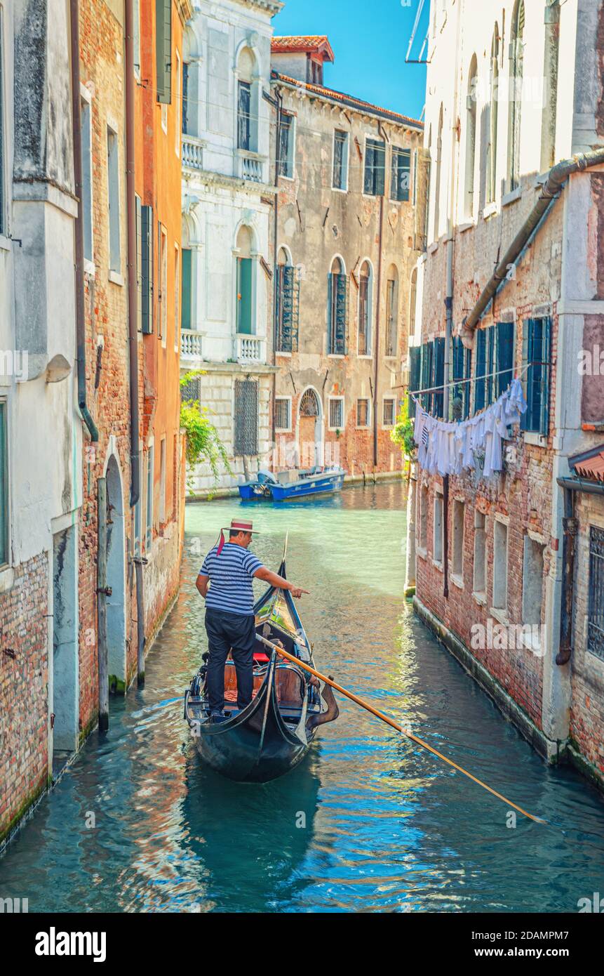 Venezia, Italia, 13 settembre 2019: Gondola a vela stretto canale tra vecchi edifici con mura di mattoni. Gondoliere vestito tradizionale bianco e blu a righe polo a maniche corte e cappello boater Foto Stock
