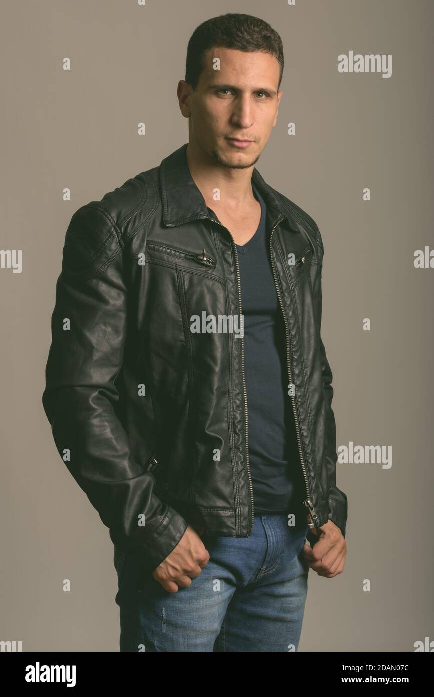 Studio di un giovane e muscoloso uomo persiano in piedi mentre indossa giacca in pelle su sfondo grigio Foto Stock