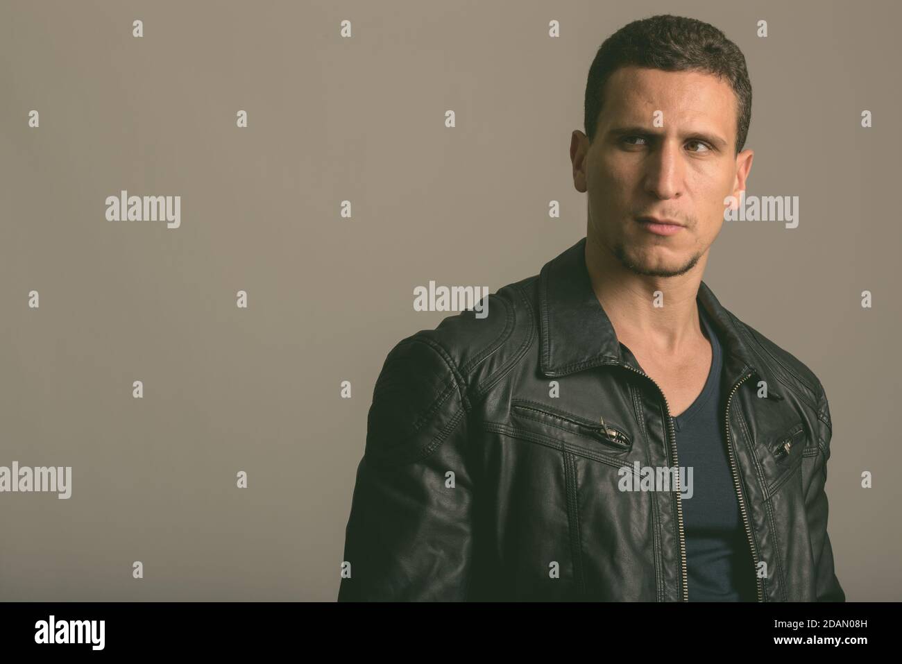 Studio di giovane muscoloso uomo persiano che pensa e guarda indossando una giacca in pelle su sfondo grigio Foto Stock