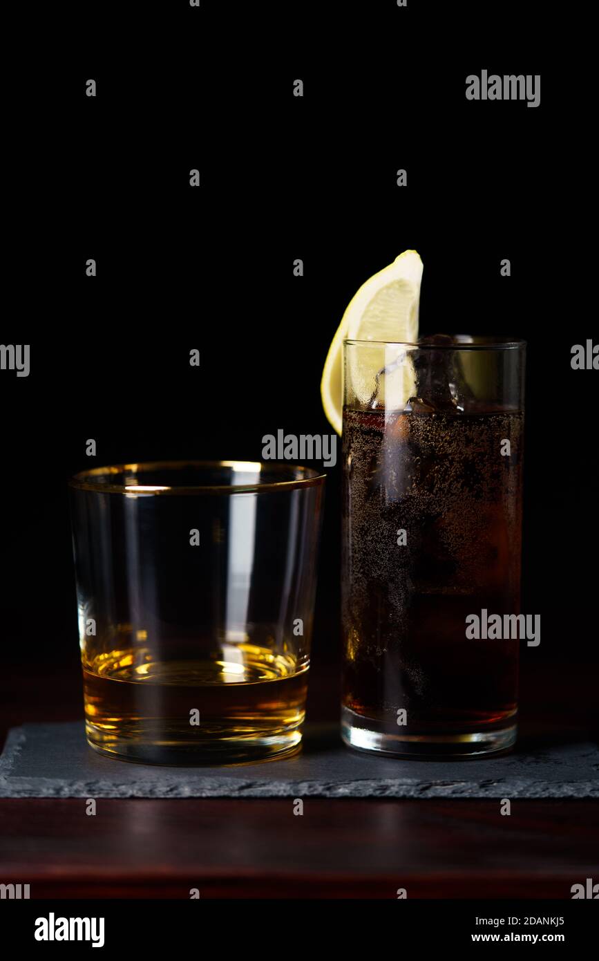 Whisky pulito e cola con ghiaccio e spicchio di limone servito su piastra di ardesia. Tavolo in legno rosso scuro, alta risoluzione Foto Stock