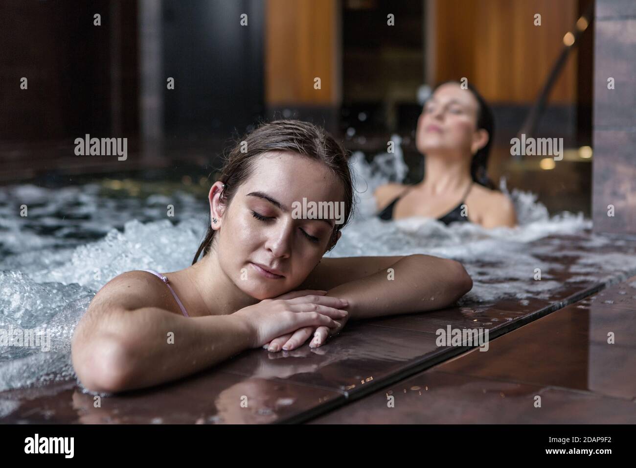 Donna tranquilla e rilassata con gli occhi chiusi che si rilassano in acqua schiumosa di piscina con idromassaggio durante il fine settimana con la ragazza nel centro termale Foto Stock