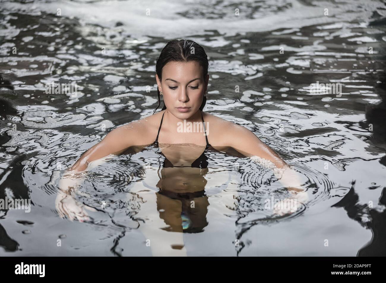 Angolo di una giovane e serena brunetta godendo di acqua di cristallo fresca in piscina mentre si ottiene la procedura di idroterapia nel centro termale Foto Stock