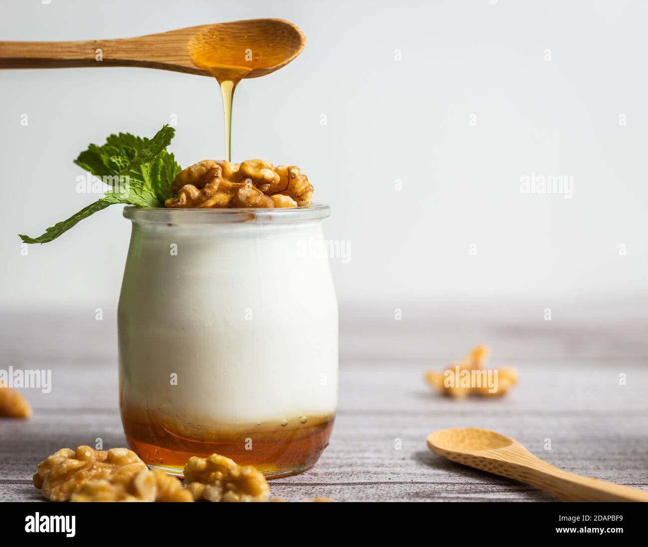 Il dessert allo yogurt con miele su un cucchiaio di legno fa cadere il miele all'interno del contenitore di yogurt con noci Foto Stock