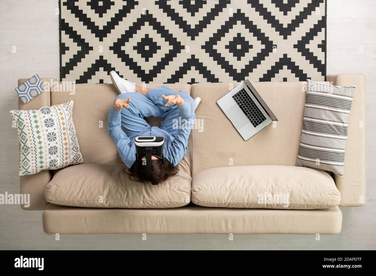Panoramica di giovane donna brunetta in pigiama blu e vr cuffie che funzionano sul divano Foto Stock