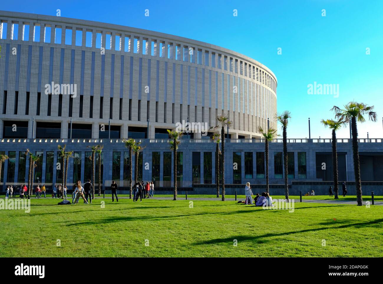 Le persone camminano nel parco mentre mantengono la 'distanza sociale' l'un l'altro. Verde prato, palme, edificio stadio Foto Stock