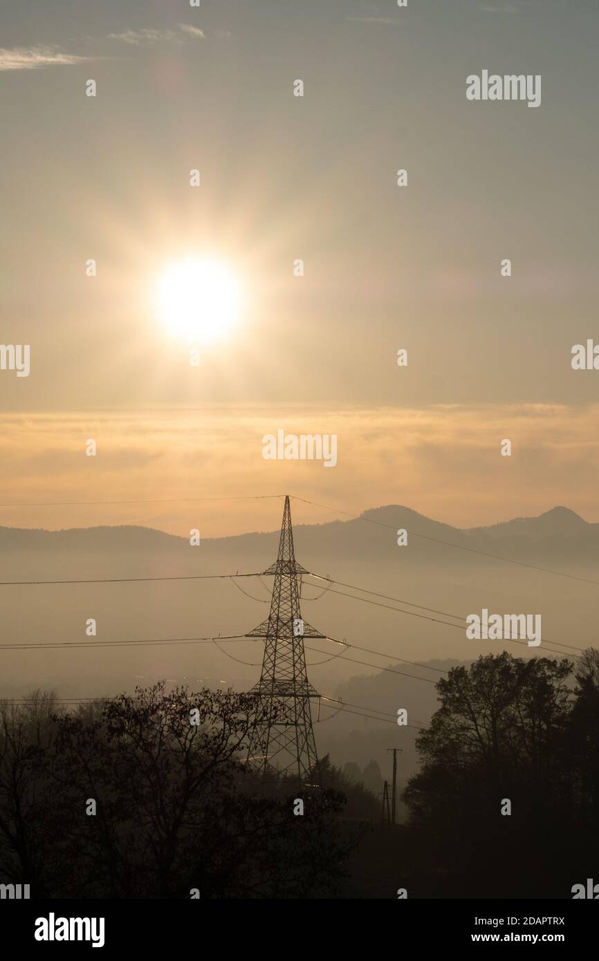 vista delle infrastrutture elettriche, dei poli elettrici e dei cavi in un bellissimo paesaggio con montagne nebbie e il sole che tramonta. Foto Stock