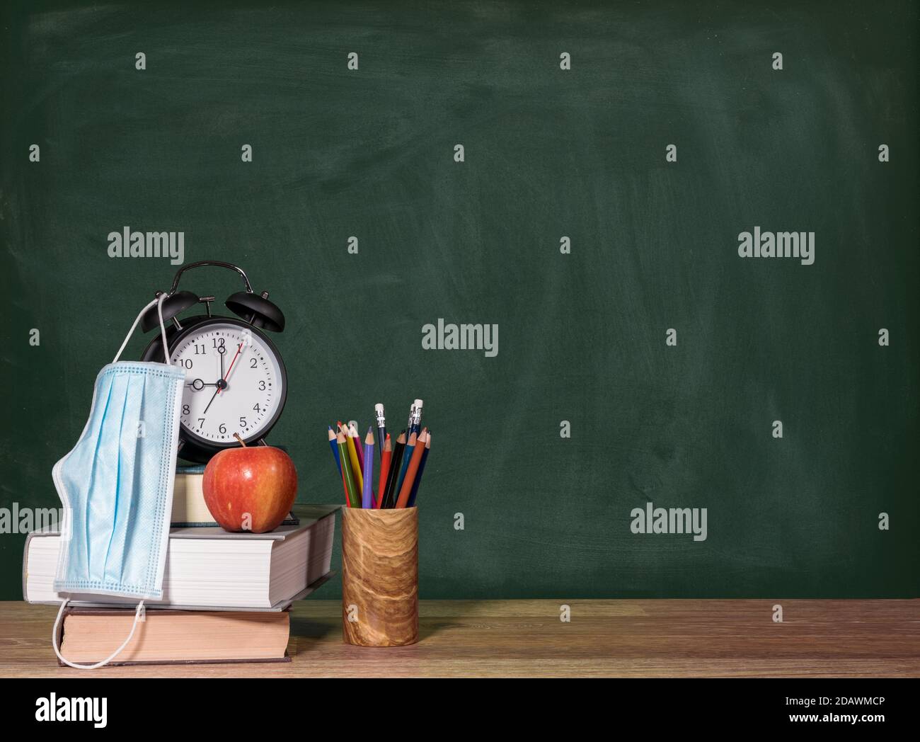 Concetto per il ritorno a scuola con coronavirus o Covid-19 con libri, mela e sveglia contro una lavagna con maschera facciale Foto Stock