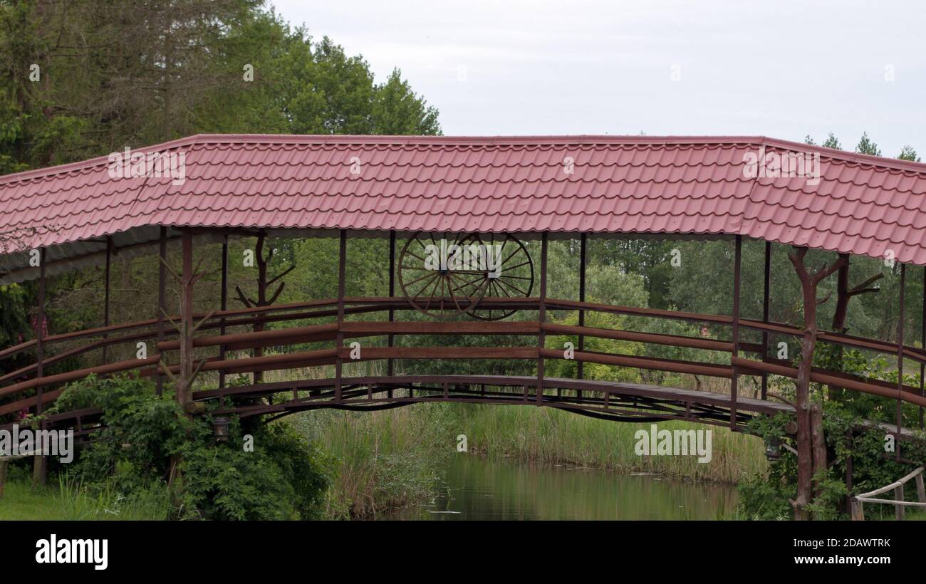 Un ponte di legno con due ruote in legno e un ringhiera con un tetto di tegole di metallo rosso scuro su a. piccolo fiume sullo sfondo di alberi verdi e cespugli Foto Stock