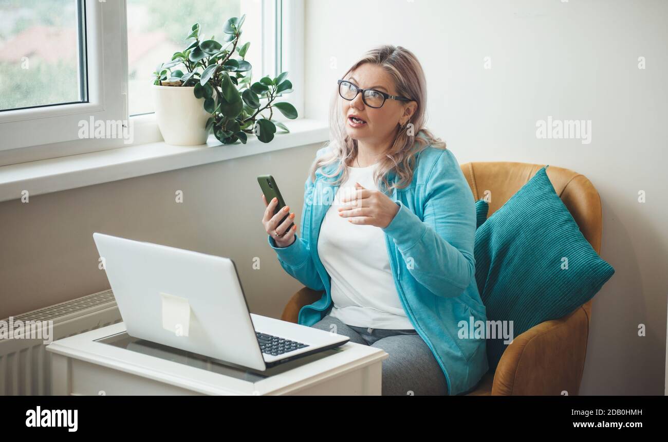 Donna anziana occupata con i capelli e gli occhiali biondi sta avendo meeting online o notebook che lavorano da casa Foto Stock