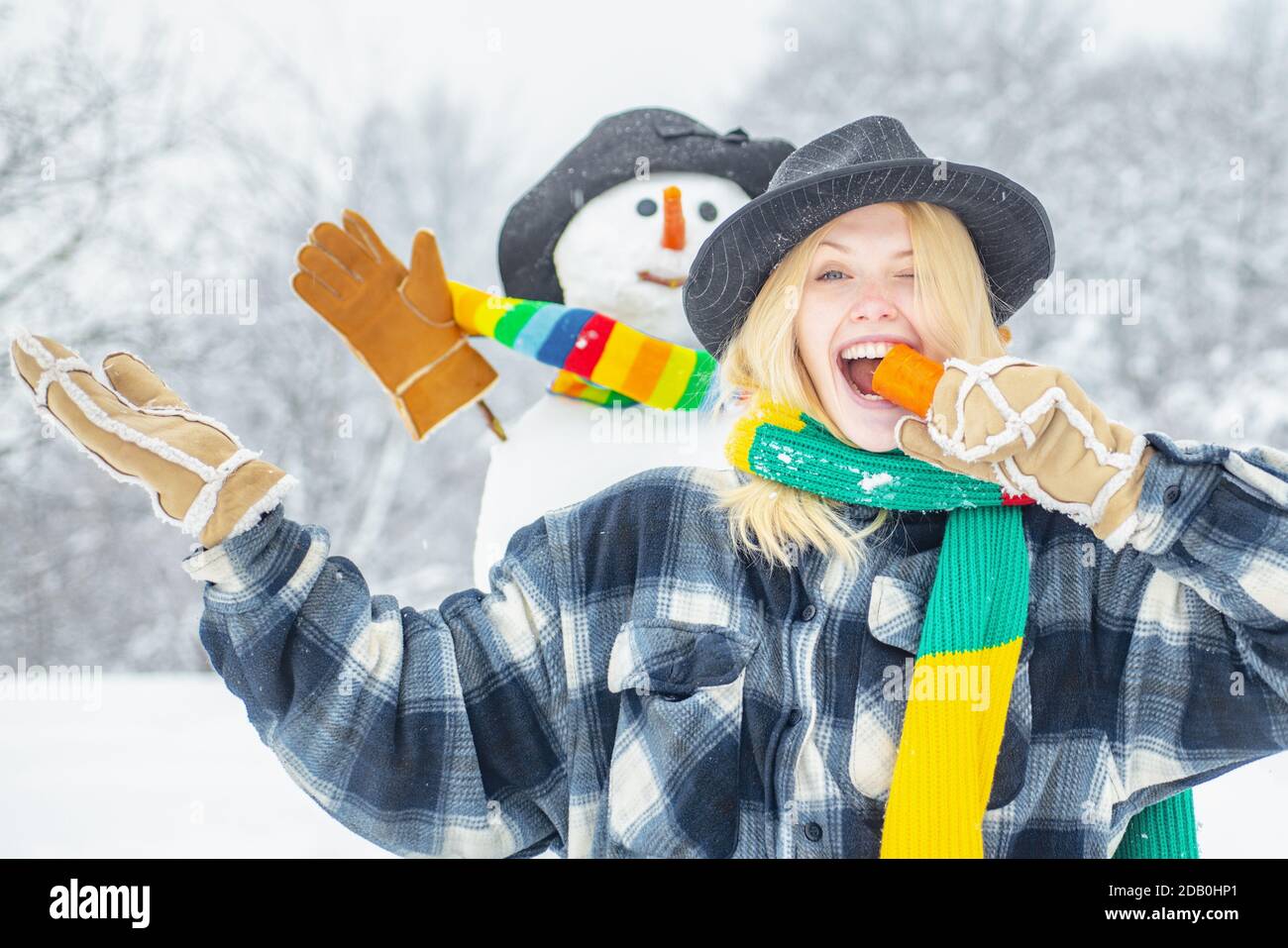 Pupazzo di neve divertente con una carota invece di un naso e in un caldo cappello lavorato a maglia su un prato nevoso su uno sfondo di neve sfocato. Innevamento uomo con nos divertenti di carote Foto Stock