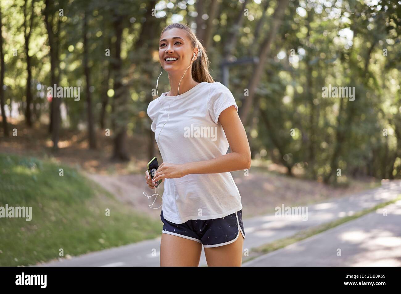 Donna ascoltare musica in esecuzione su asfalto pista estate parco caucasico Auricolari donne jogging mattina soleggiato Runner allenamento tenere smartphone in Healt. Mano Foto Stock
