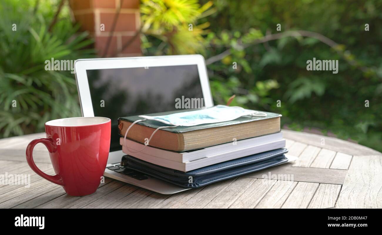 Maschera sulla parte superiore dei libri impilati e computer portatile aperto con una tazza rossa. Studio a casa durante il concetto pandemico. Foto Stock