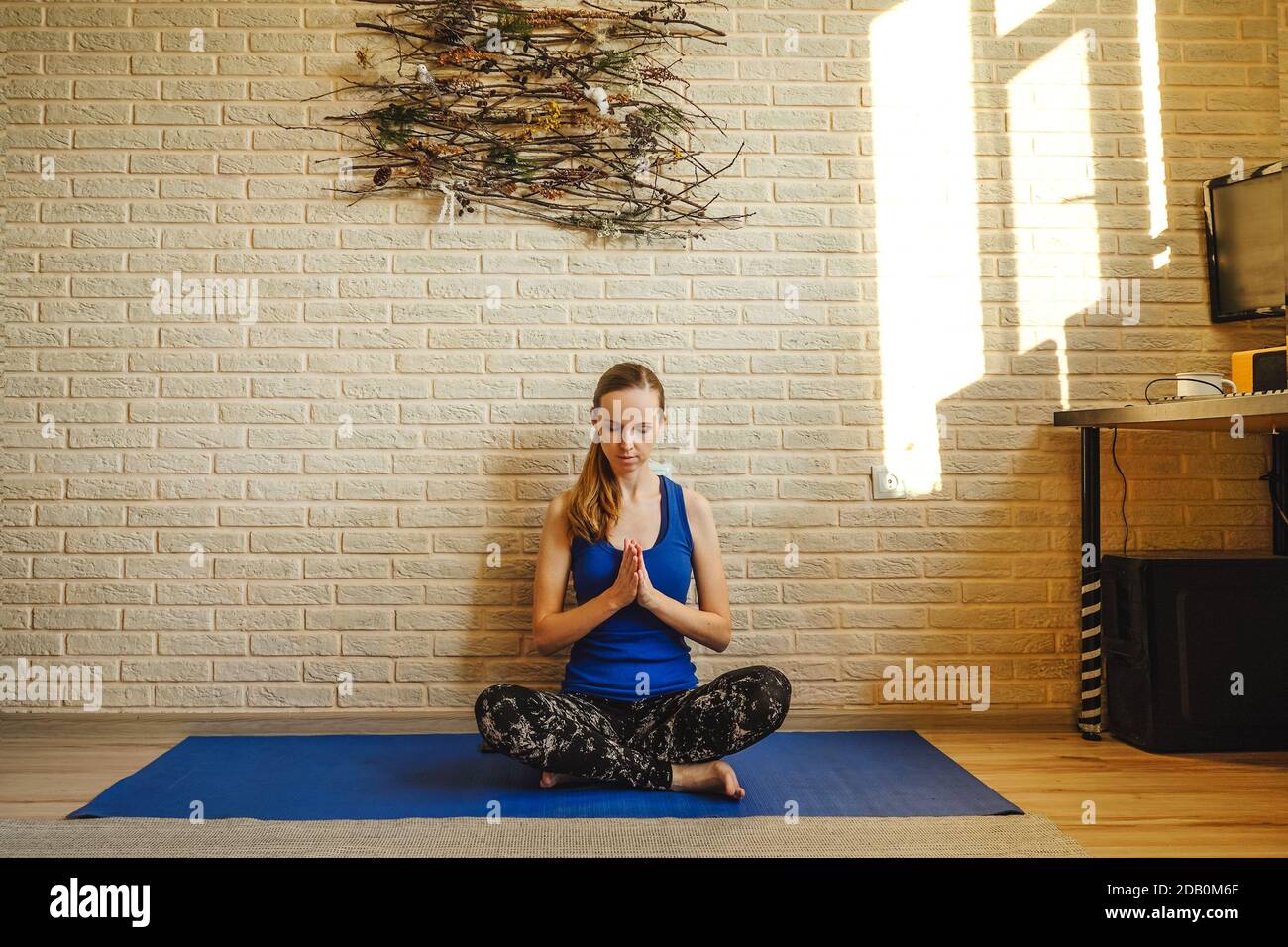 Donna che fa yoga a casa. Tappetino e camicia blu. Namaste, giorno di sole. Pannelli naturali sulla parete in mattoni, design sostenibile. Assistere all'healt mentale Foto Stock