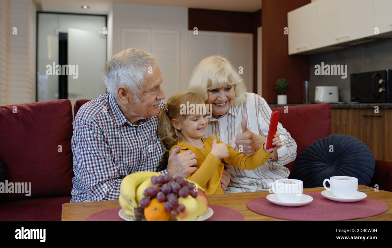 Coppia senior nonni con bambina nipotina passare il tempo a casa insieme, seduto sul divano in soggiorno, utilizzando il telefono cellulare. Giocare, fare foto selfie insieme sullo smartphone Foto Stock