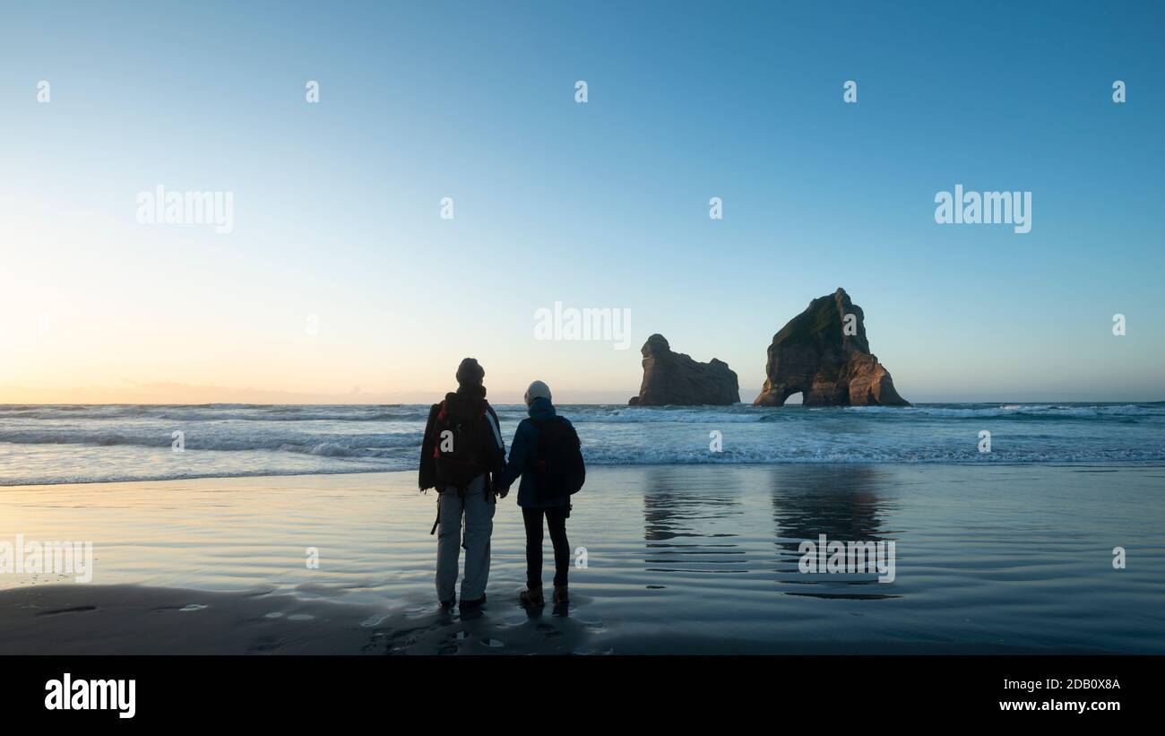 Coppia che si trova sulla spiaggia di Wharariki e guarda l'Isola di Archway al tramonto, Isola del Sud della Nuova Zelanda Foto Stock