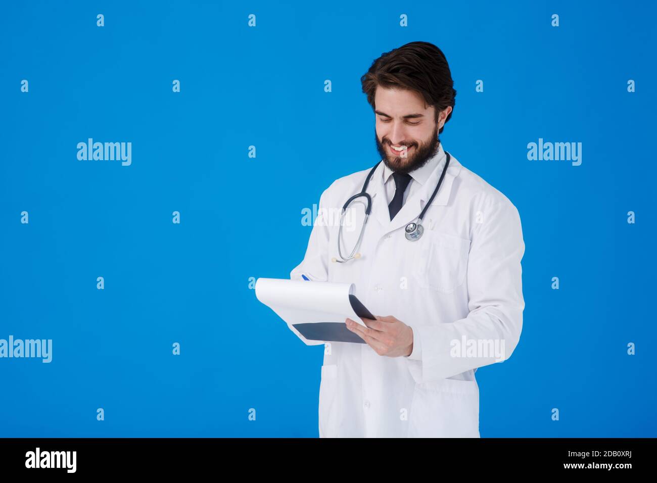 un giovane medico con la bearded in un cappotto medico bianco sopra uno sfondo blu scrive i reclami del paziente su un tablet con documenti e medici Foto Stock