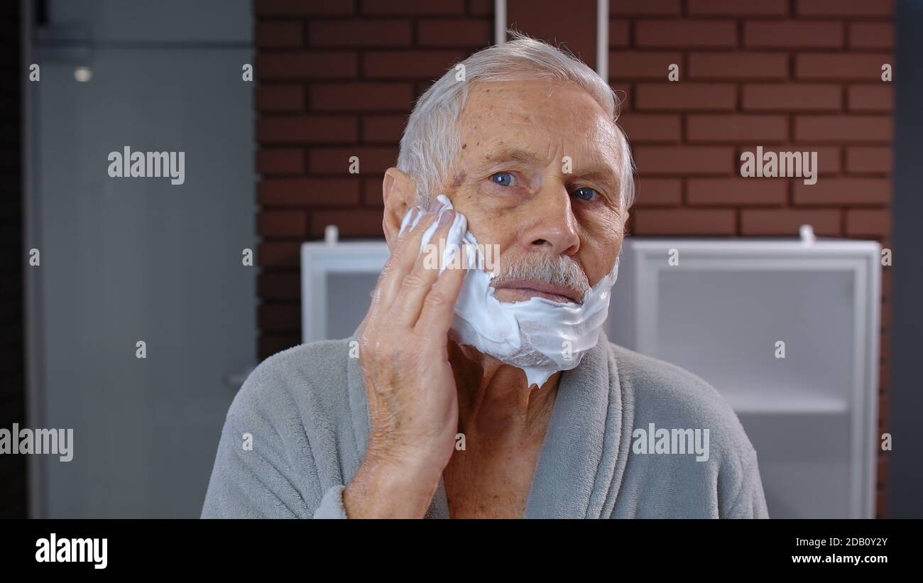 Nonno anziano attraente in accappatoio che applica la schiuma al viso per  la rasatura e guardare nello specchio. Bel nonno anziano che fa l'igiene  della pelle del mattino dopo la doccia a