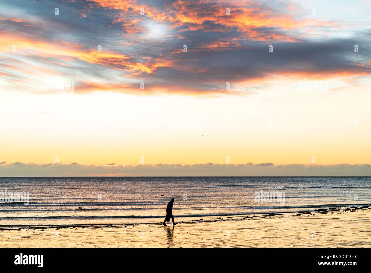 Uomo che cammina sulla costa portando una borsa sotto il cielo dell'alba  sul canale della Manica, visto dalla spiaggia di Viking Bay nella località  balneare di Broadstairs, Kent. Bassa marea, sabbia bagnata