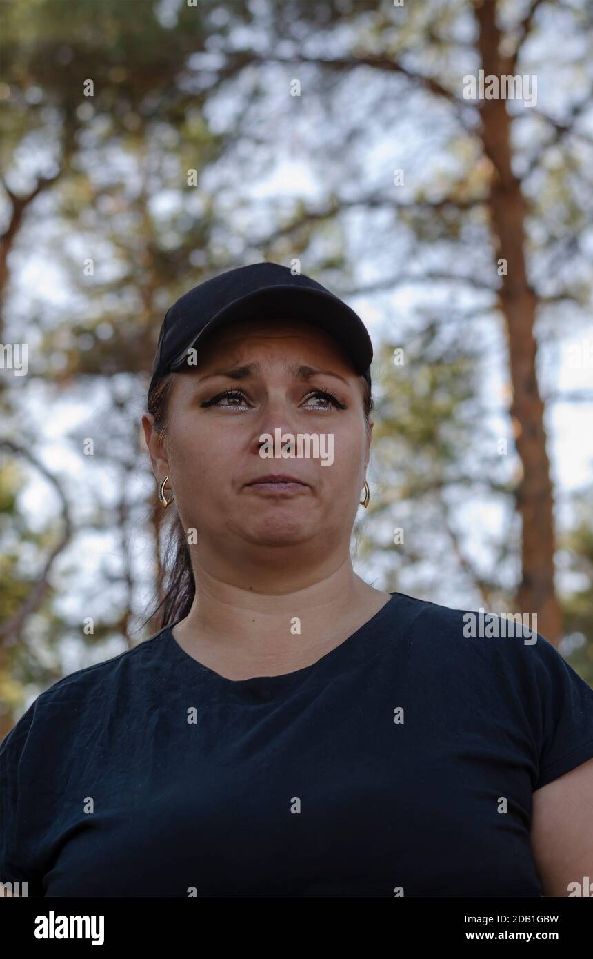 Una donna armena di 40 anni guarda lontano con le labbra strette. Ritratto di una donna in un cappellino da baseball nero e camicia Foto Stock