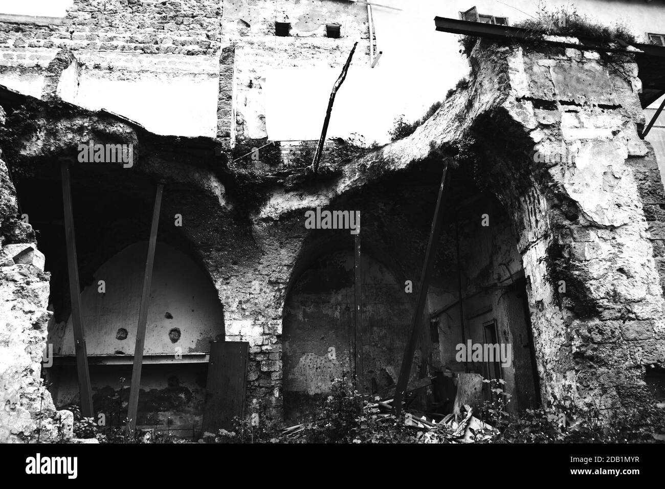 Casa abbandonata in rovina vicino a un edificio residenziale. Muro di pietra rotto e archi sostenuti da arbusti di metallo. Ridurre i concetti di pericolo. Foto Stock