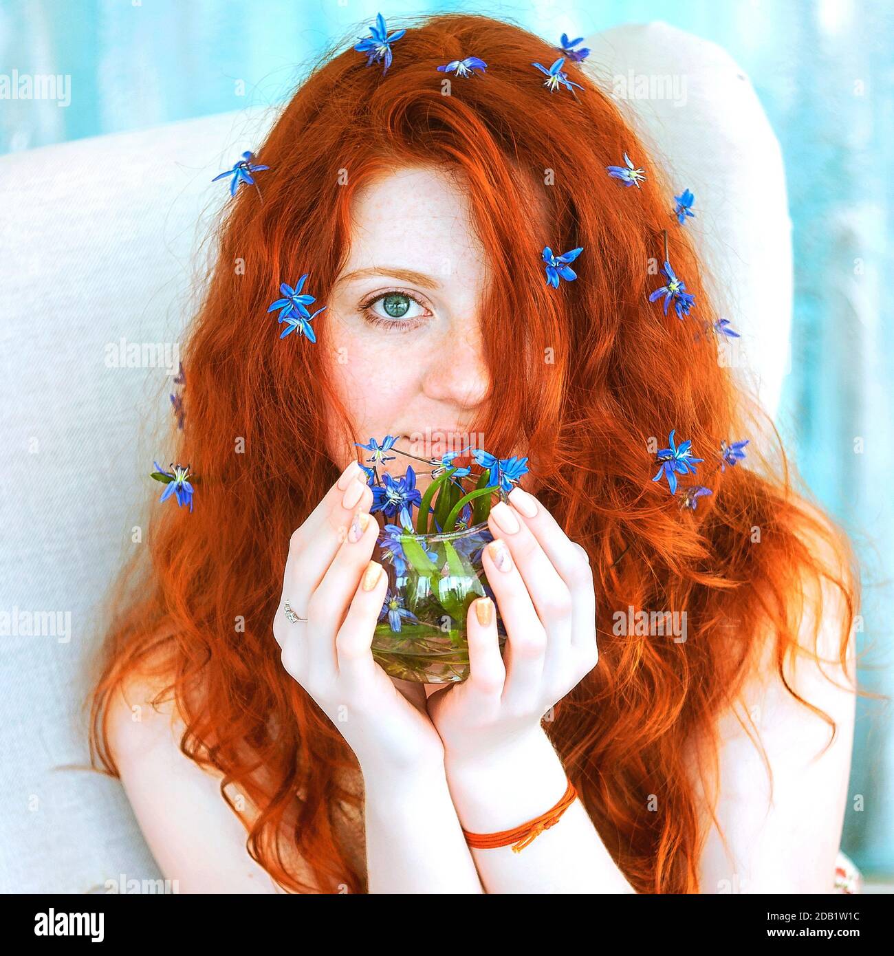 Una ragazza carina di testa rossa è seduta in una grande sedia con un bouquet di gocce di neve blu selvatiche nelle sue mani. Foto Stock