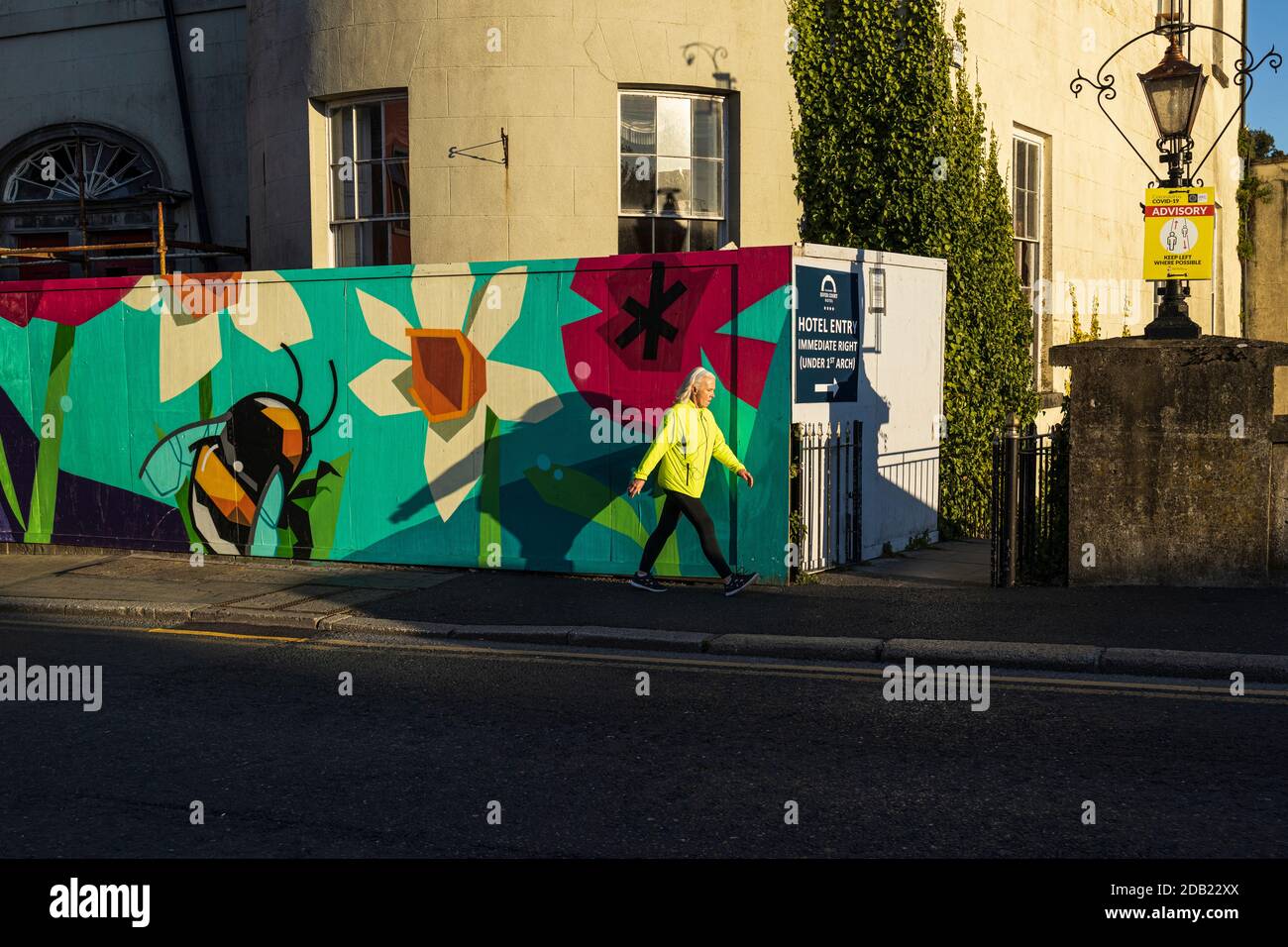 Donna anziana in giacca ad alta visibilità Power Walking sul Il marciapiede passa davanti a un'accavallamento dipinto presso il River Court Hotel E un cartello consultivo Covid 19 in K Foto Stock