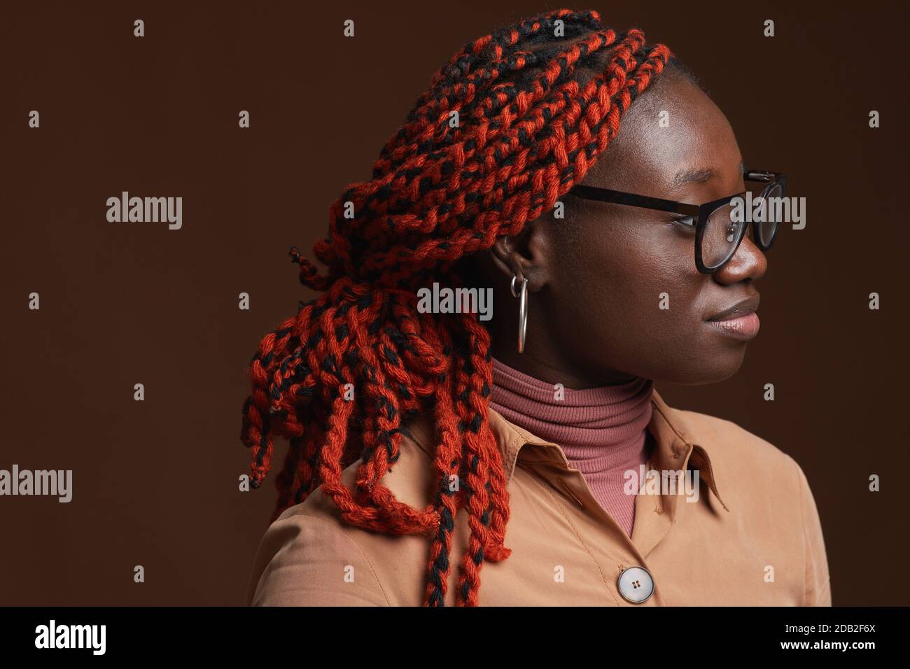 Testa e spalle Vista laterale ritratto di donna afroamericana contemporanea con capelli intrecciati che si guardano via mentre si posa su sfondo marrone scuro in studio, copia spazio Foto Stock