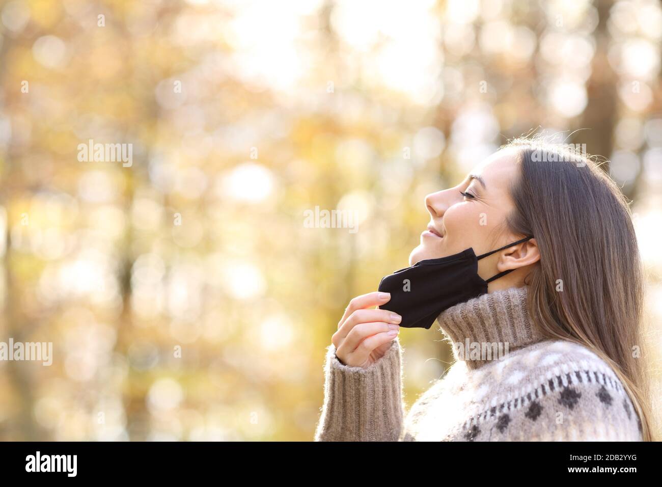 Vista laterale ritratto di una donna soddisfatta che allevia il decollo maschera protettiva per respirare aria fresca nella stagione autunnale un parco Foto Stock