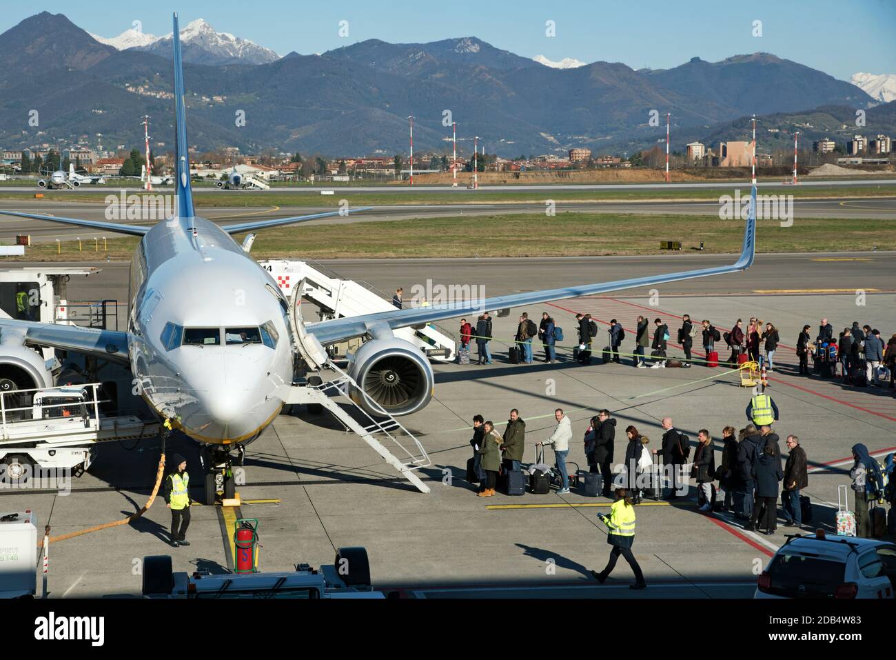 I passeggeri in fila per salire a bordo di un jet Aircraft in aeroporto il giorno di sole, visto dalla finestra del terminal. Paesaggio di montagna è sullo sfondo Foto Stock