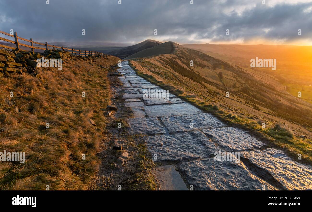 Sentiero in pietra che conduce a una lunga dorsale di montagna MAM Tor con nubi tempesta e luce dorata. Foto Stock