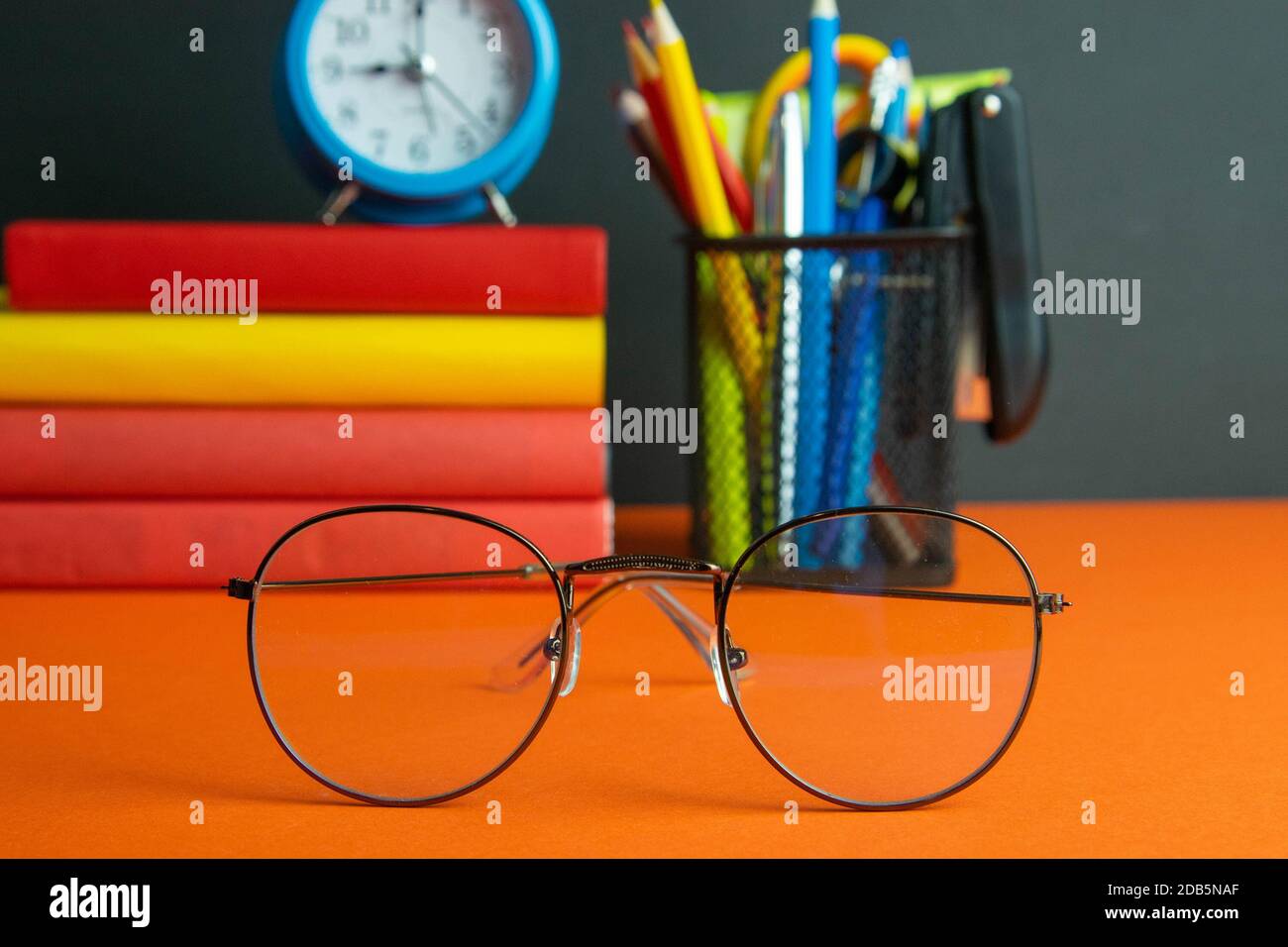 Una pila di libri, bicchieri da donna in primo piano, orologio blu e un bicchiere di matite si trovano su uno sfondo arancione. Foto Stock