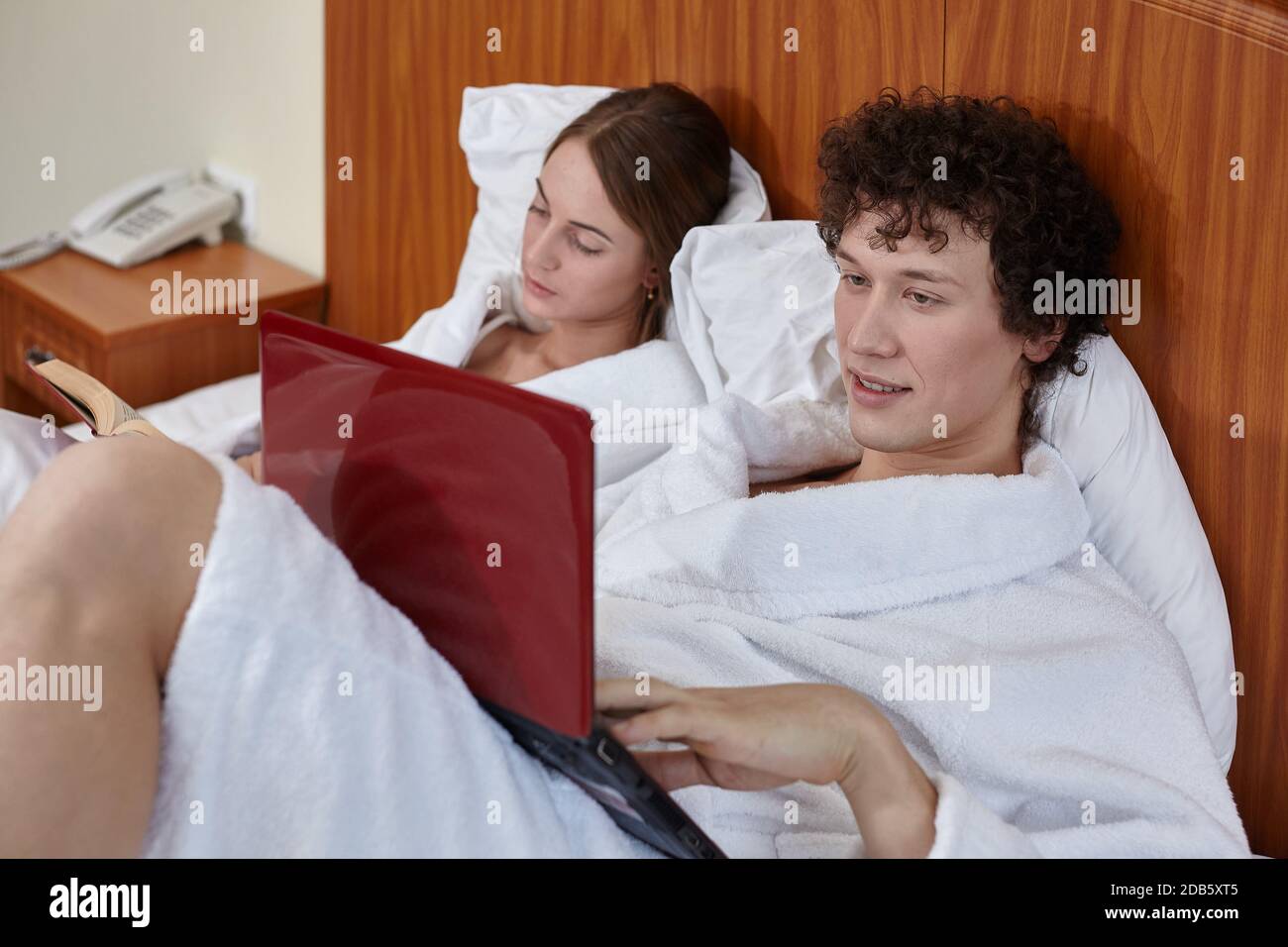 La famiglia giovane con il computer portatile e il libro si trova sul letto in camera a casa. Fine settimana o periodo di isolamento, distanza sociale. Apprendimento e lavoro a distanza. Felice yo Foto Stock