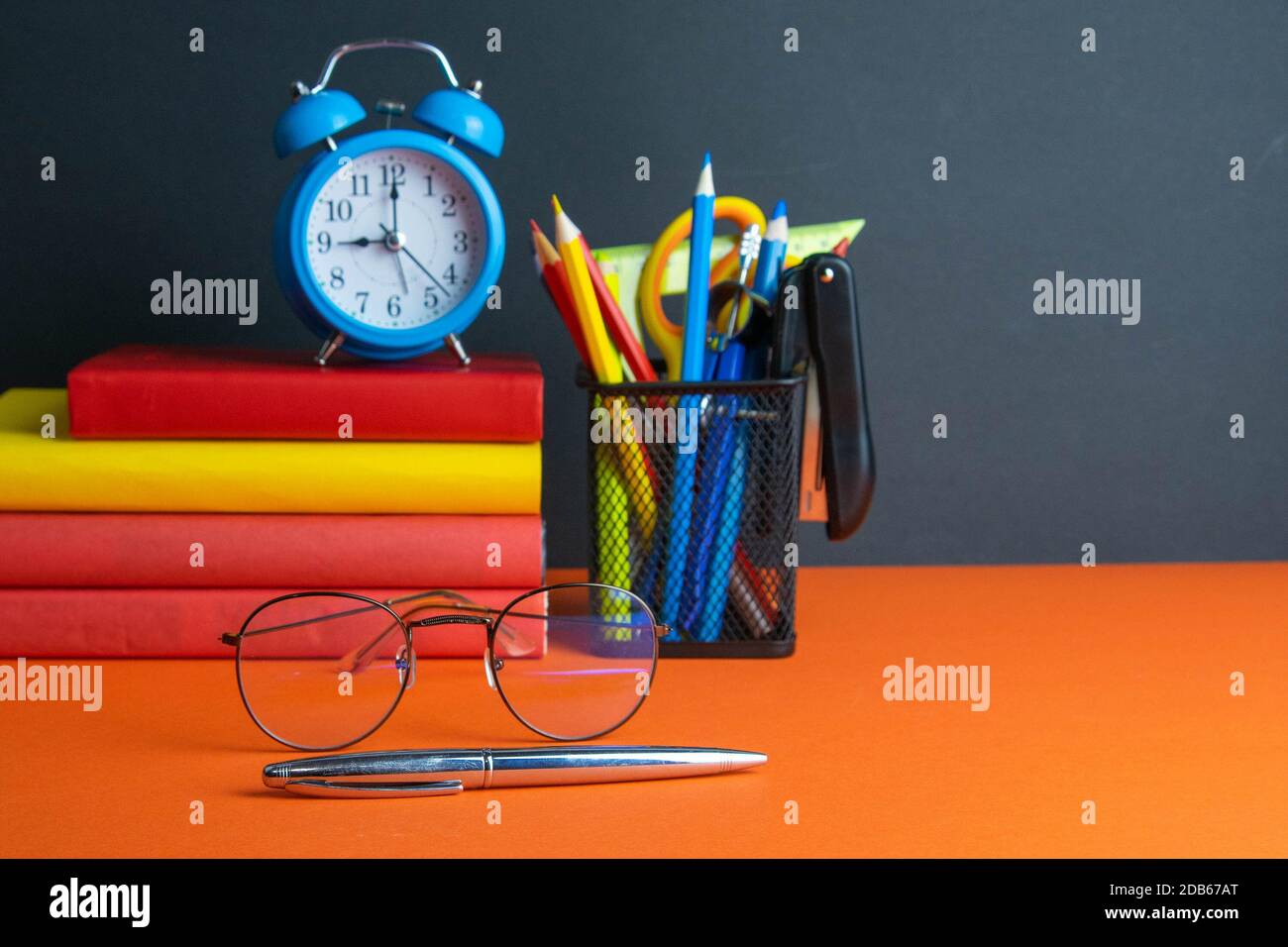 Una pila di libri, bicchieri da donna in primo piano, orologio blu e un bicchiere di matite si trovano su uno sfondo arancione. Foto Stock