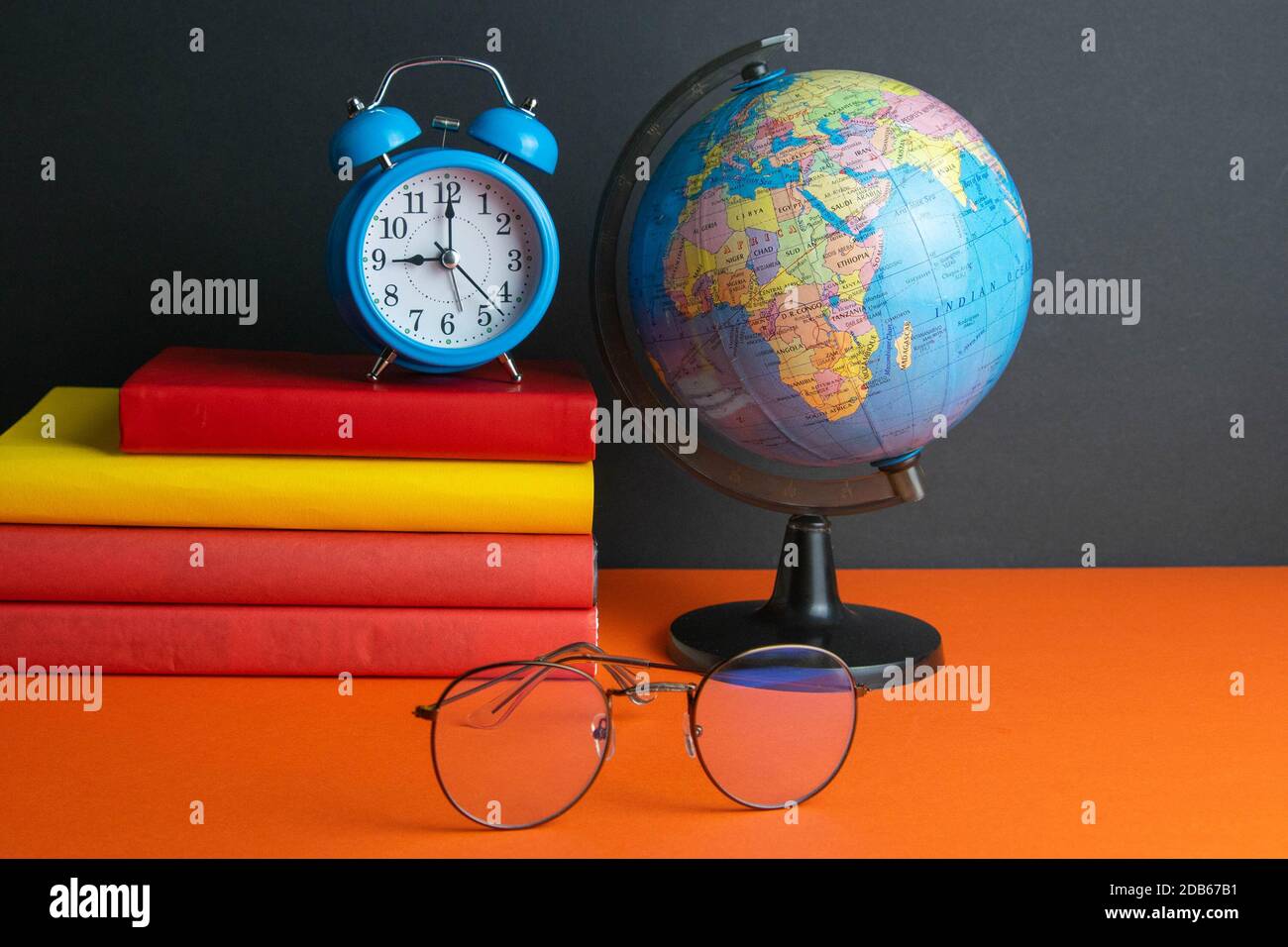 Una pila di libri, orologio, globo mondiale e occhiali da donna in primo piano, si trovano su uno sfondo arancione. Foto Stock