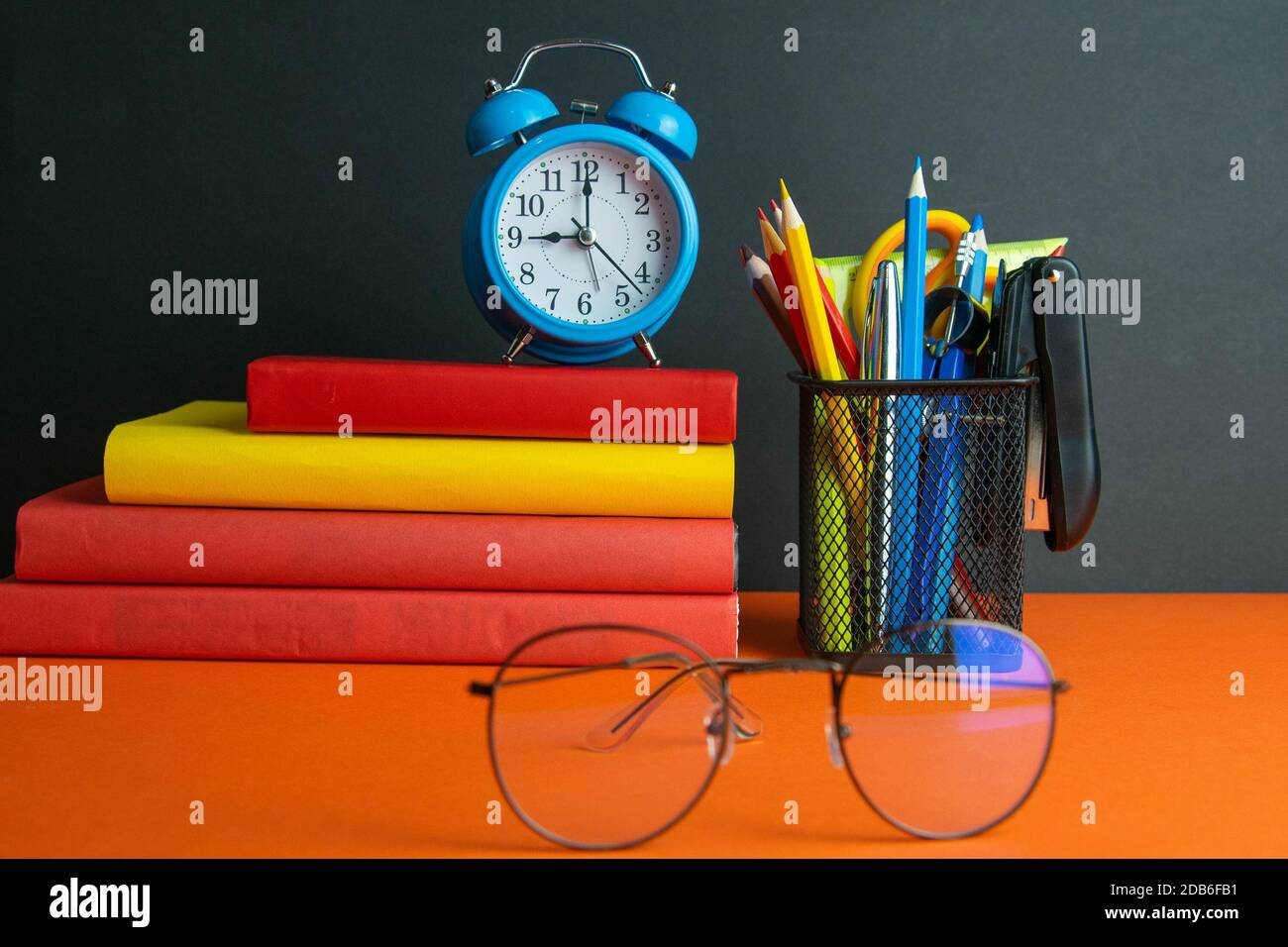 Una pila di libri, bicchieri da donna in primo piano, orologio blu e un bicchiere di matite si trovano su uno sfondo arancione. Foto Stock