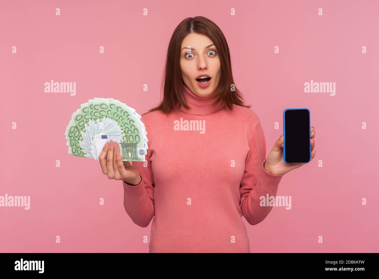 Donna bruna estremamente sorpresa in felpa rosa che tiene smartphone con schermo vuoto e fan di euro guardando fotocamera con espressione scioccata. Poll Foto Stock