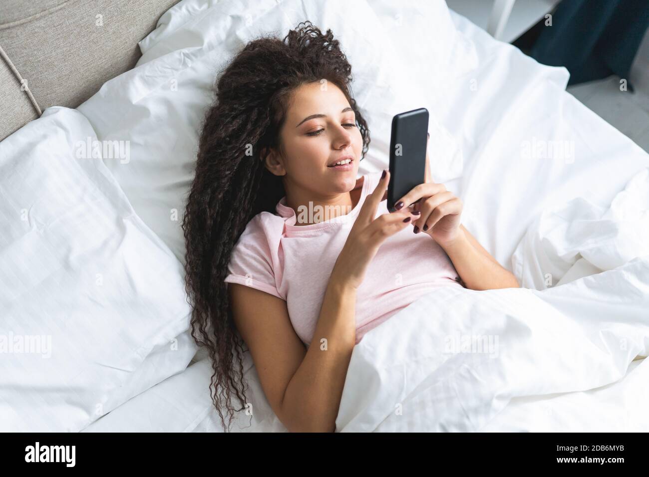 La giovane donna sorridente dei capelli si trova nel letto e usa lo smartphone. Dipendenza dai social media, datazione online. Foto Stock