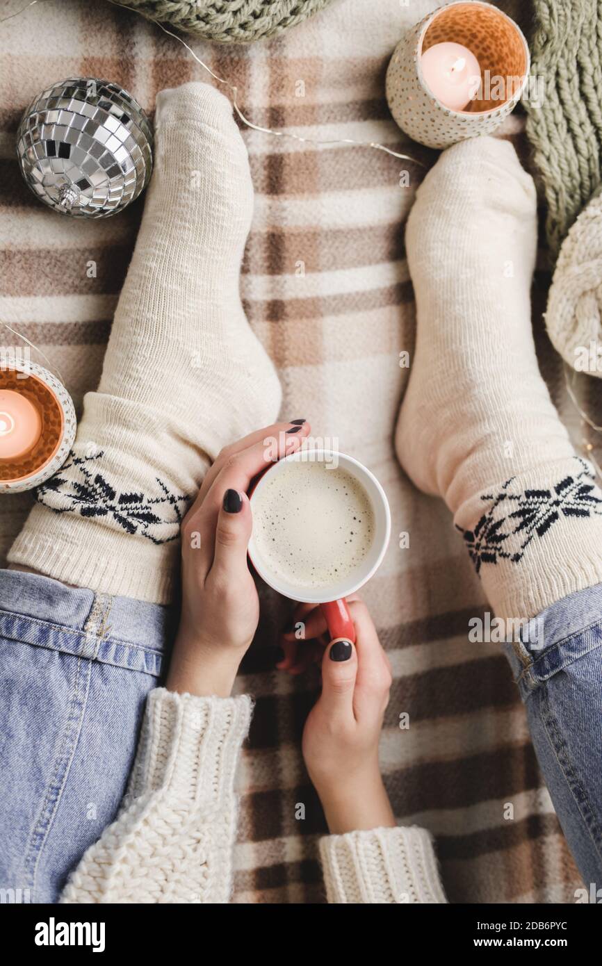 La giovane donna si siede sul plaid in un maglione bianco di lana e le calze di maglia tiene la tazza di cacao nelle sue mani. Higge Capodanno, Natale accogliente, preparazione Foto Stock