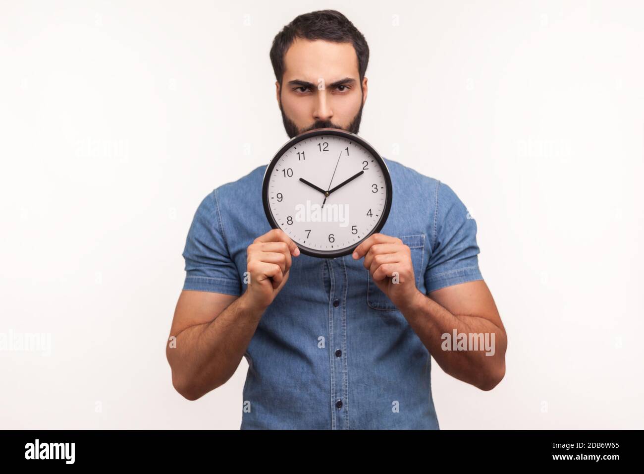 Uomo serio e bossy con la barba che tiene in mano grande orologio da parete guardando la fotocamera con dispiaciuto, tempo di agire, conto alla rovescia. Studio al coperto isolato Foto Stock
