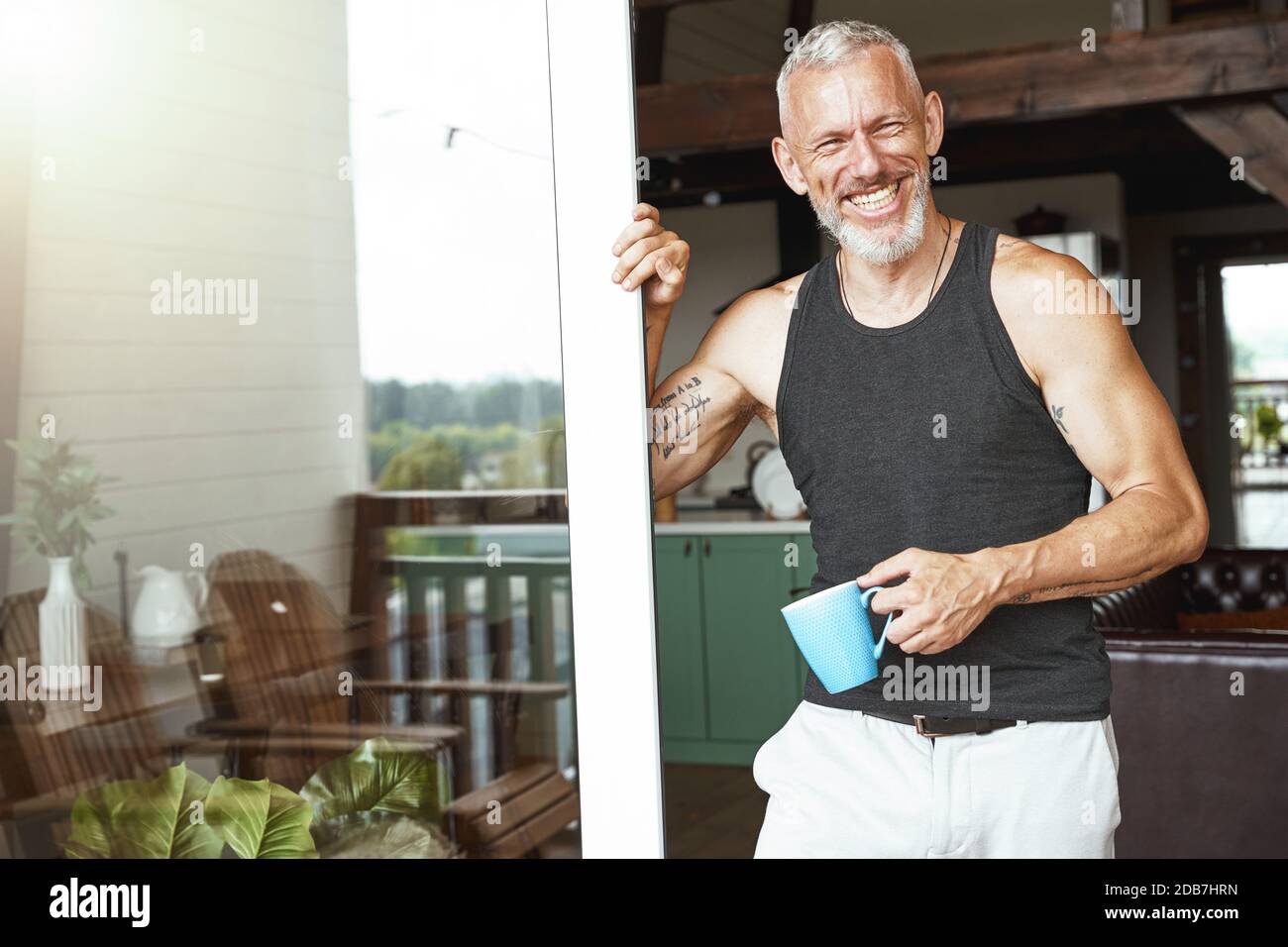 Mattina perfetta. Bell'uomo caucasico di mezza età positivo con caffè in piedi vicino all'ingresso terrazza del suo appartamento moderno Foto Stock