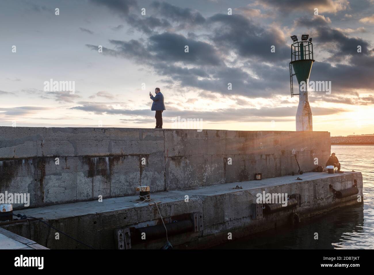 Uomo di mezza età che indossa una maschera facciale, scattando foto selfie sul molo del porto durante il tramonto, Burgas Harbour, Bulgaria Foto Stock