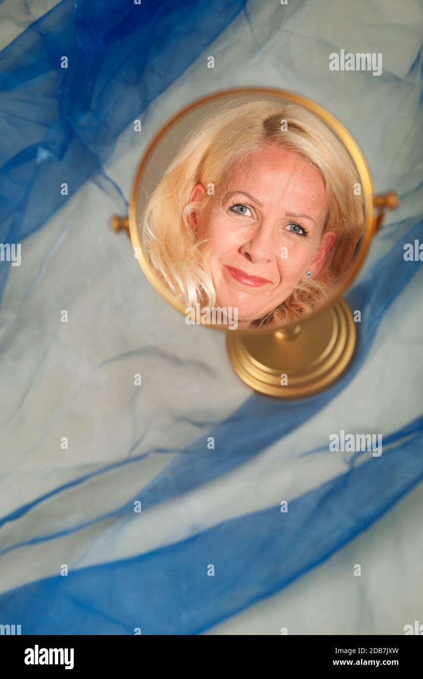 Faccia di una donna bionda di mezza età in uno specchio rotondo del trucco contro uno sfondo blu del tessuto nel formato verticale. Foto Stock
