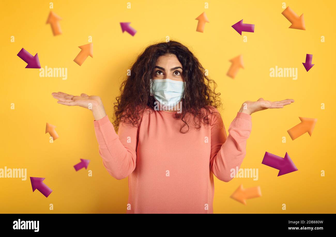 Ragazza con maschera facciale è confusa quale freccia seguire. Concetto di dubbio sulle leggi che disciplinano il covid-19. Sfondo giallo Foto Stock